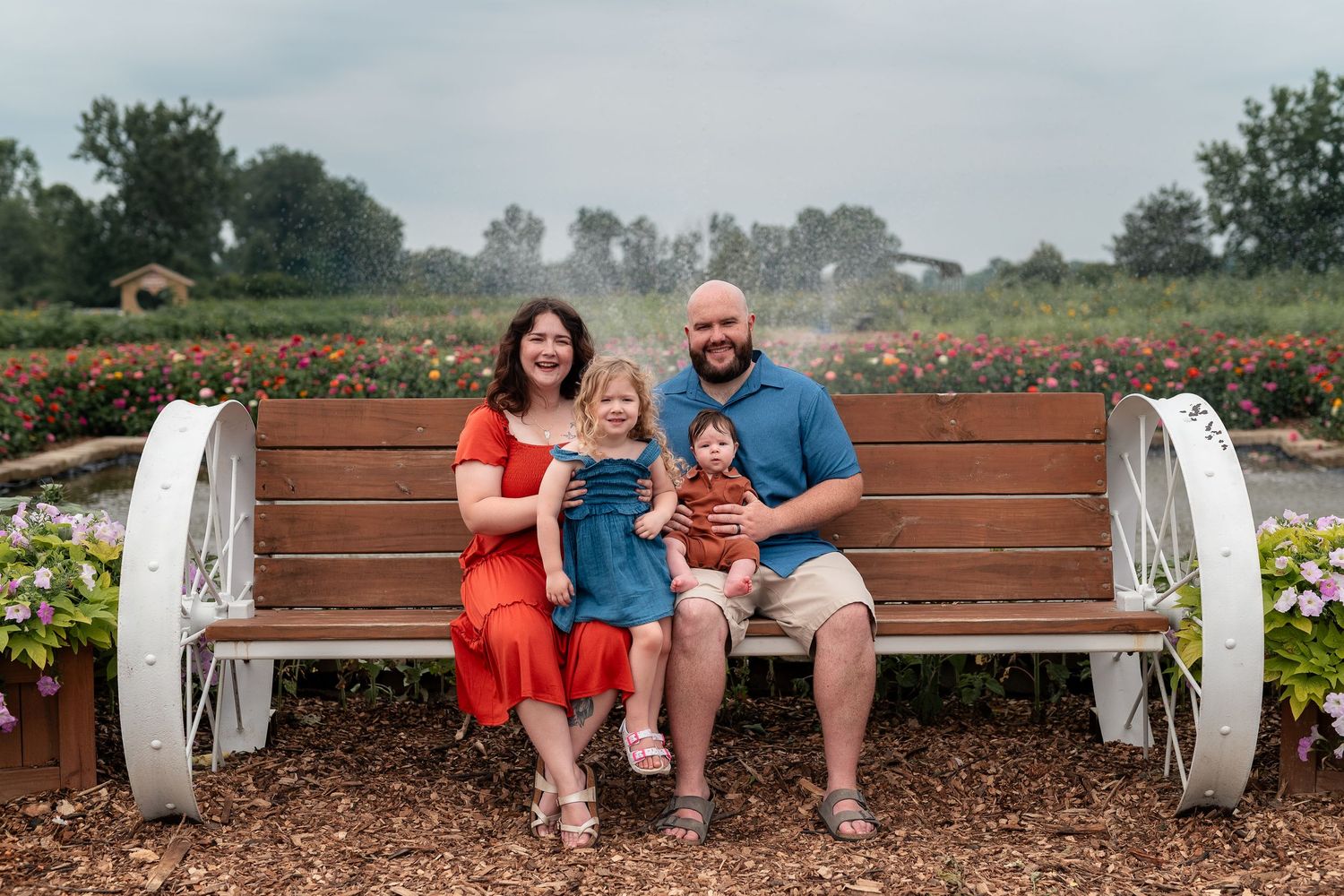 Family of four smiling on a wooden bench surrounded by colorful flowers at an outdoor farm.