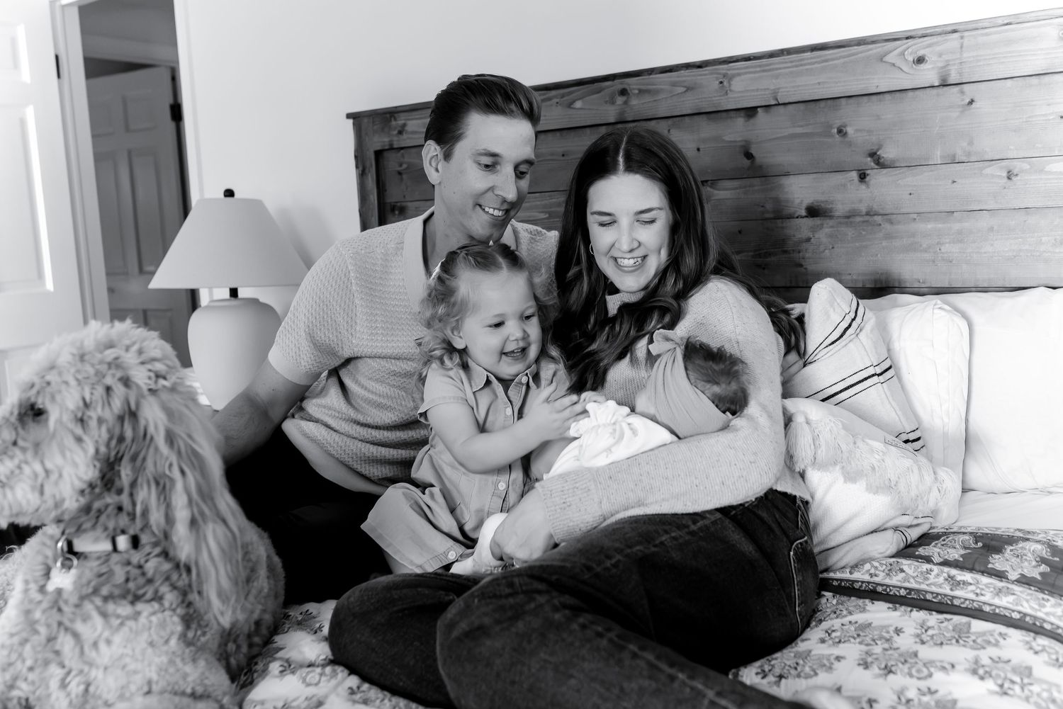 Black and white photo of happy family with newborn baby and toddler sitting on bed with dog.