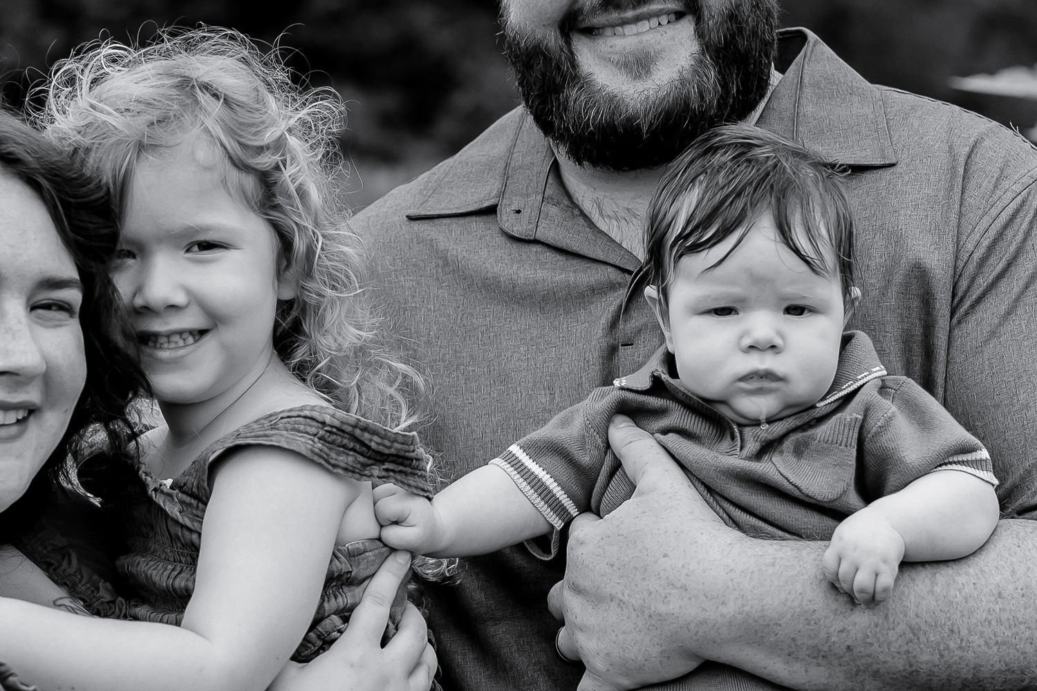 Black and white family portrait of parents with young curly-haired girl and baby boy outdoors.