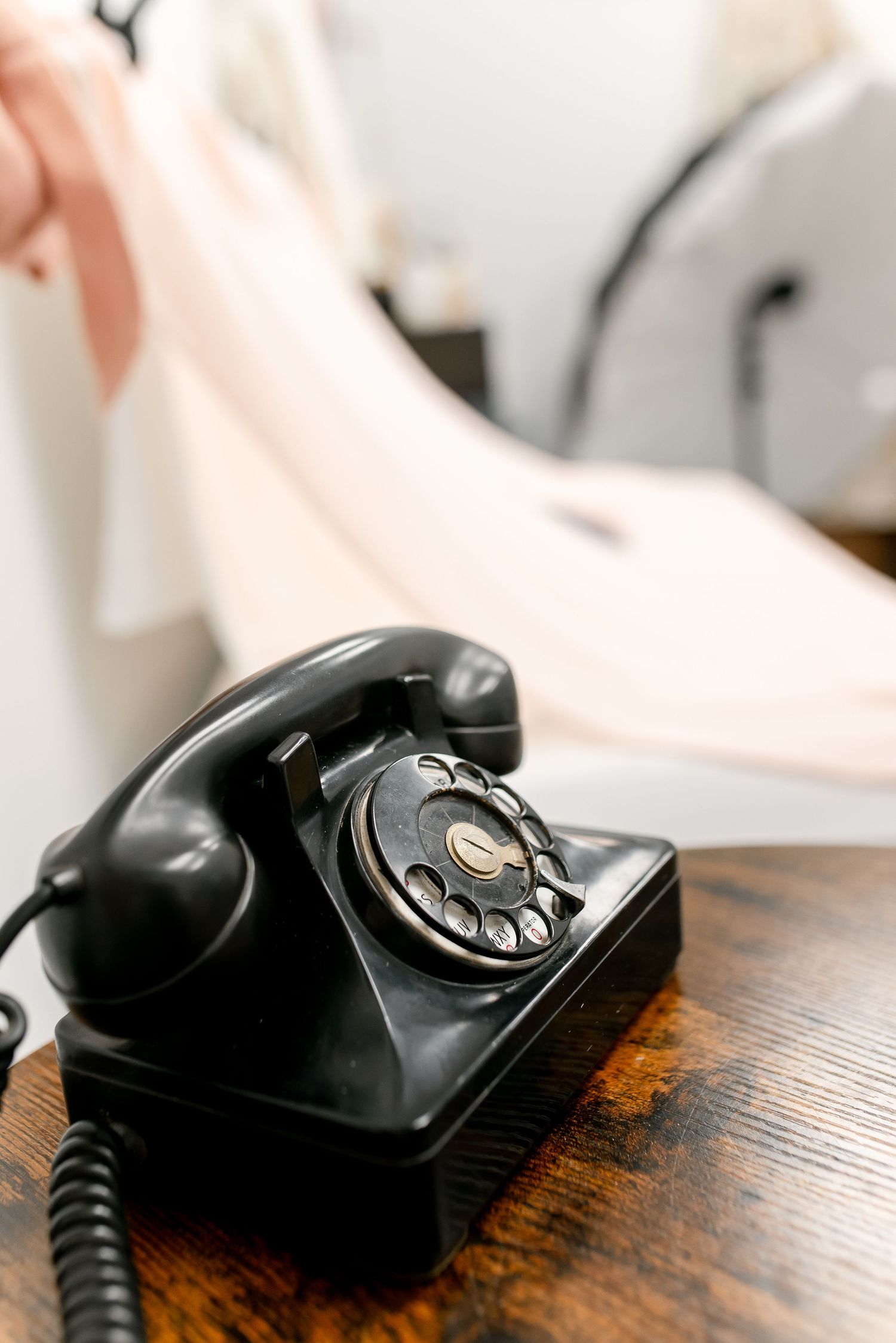 A vintage black rotary telephone sits on a wooden table in a softly lit room with blurred background.