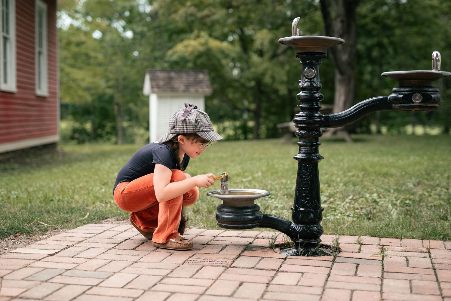 Child crouching by a vintage black cast iron water pump fountain on a brick path in a green park.