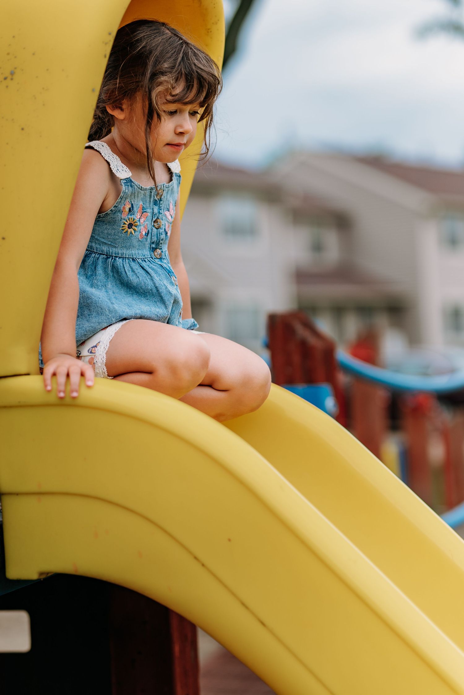 A child in denim clothing sits on a bright yellow curved playground structure on a sunny day.