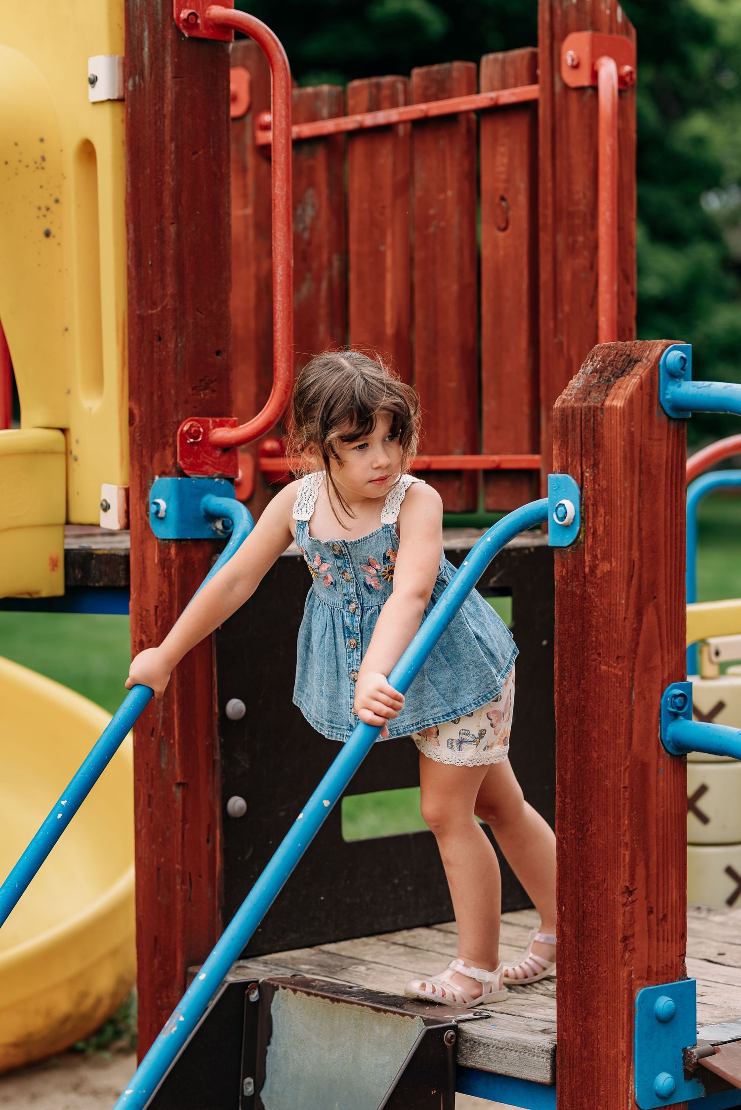 A young child in a denim dress playing on a colorful outdoor playground structure.