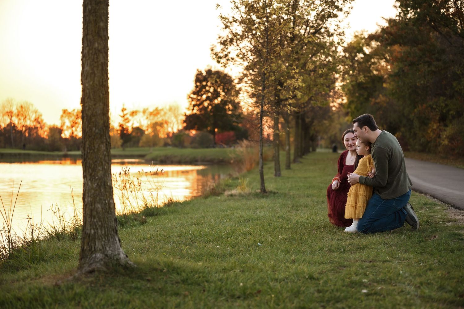 A couple sits on a grassy hill during golden hour as the sun sets over a tranquil lake in the background.