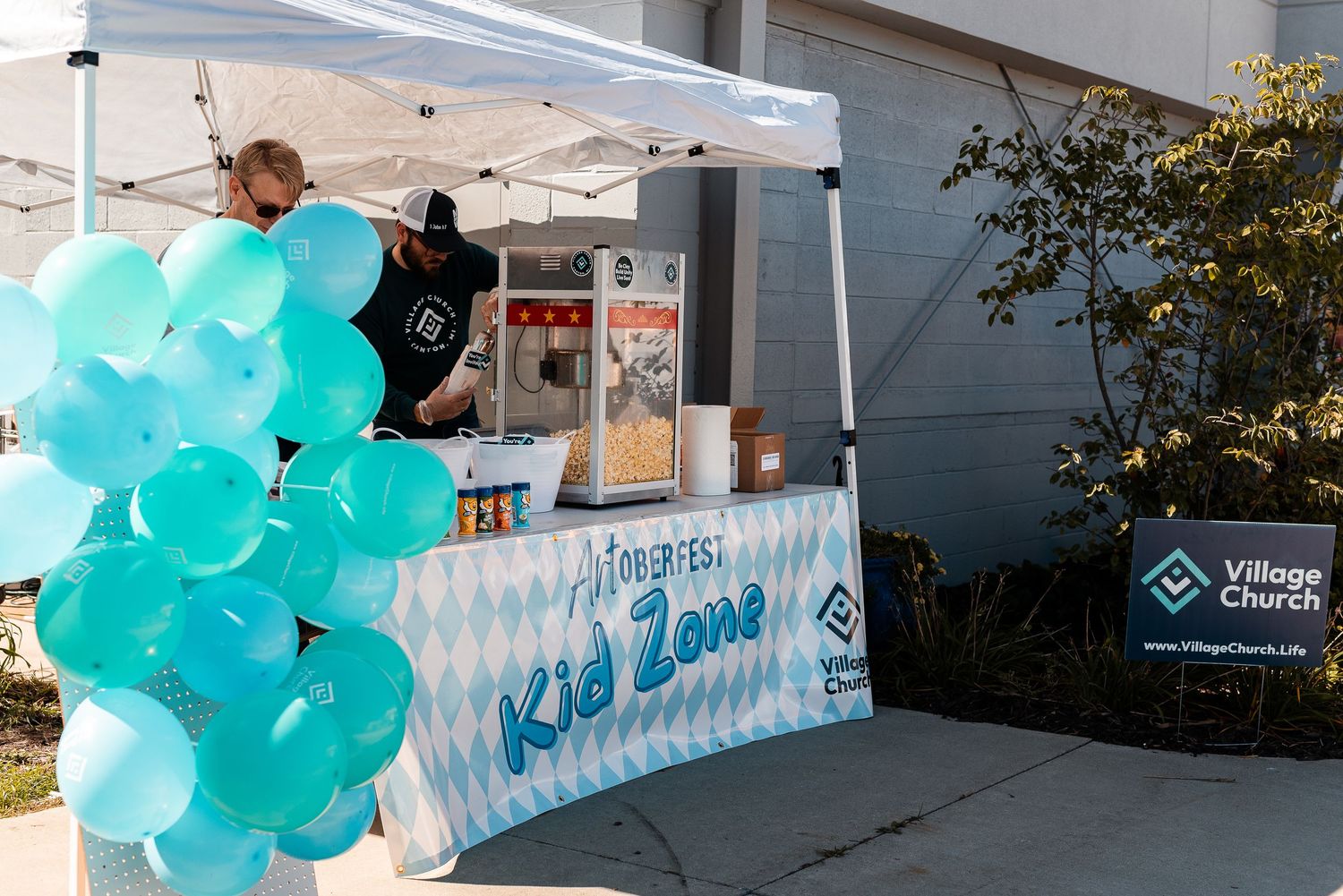 Man sets up a Kid Zone booth with teal balloons at Village Church outdoor event under a white canopy tent.