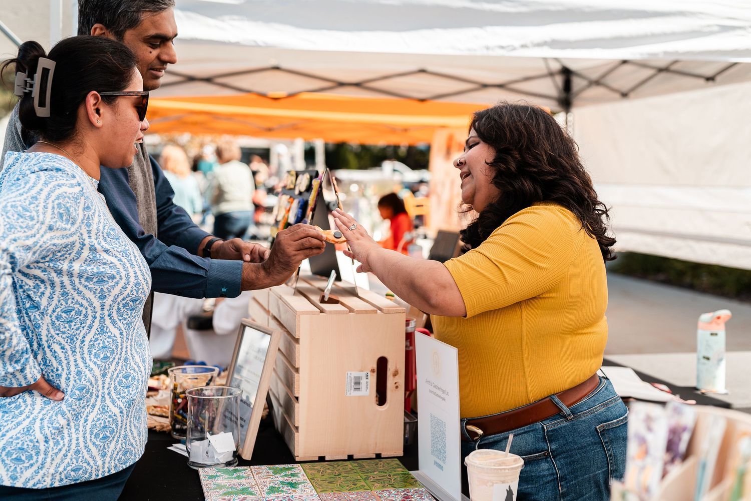 Vendor in yellow shirt handing a bottle to customers at an outdoor market booth under a white tent.