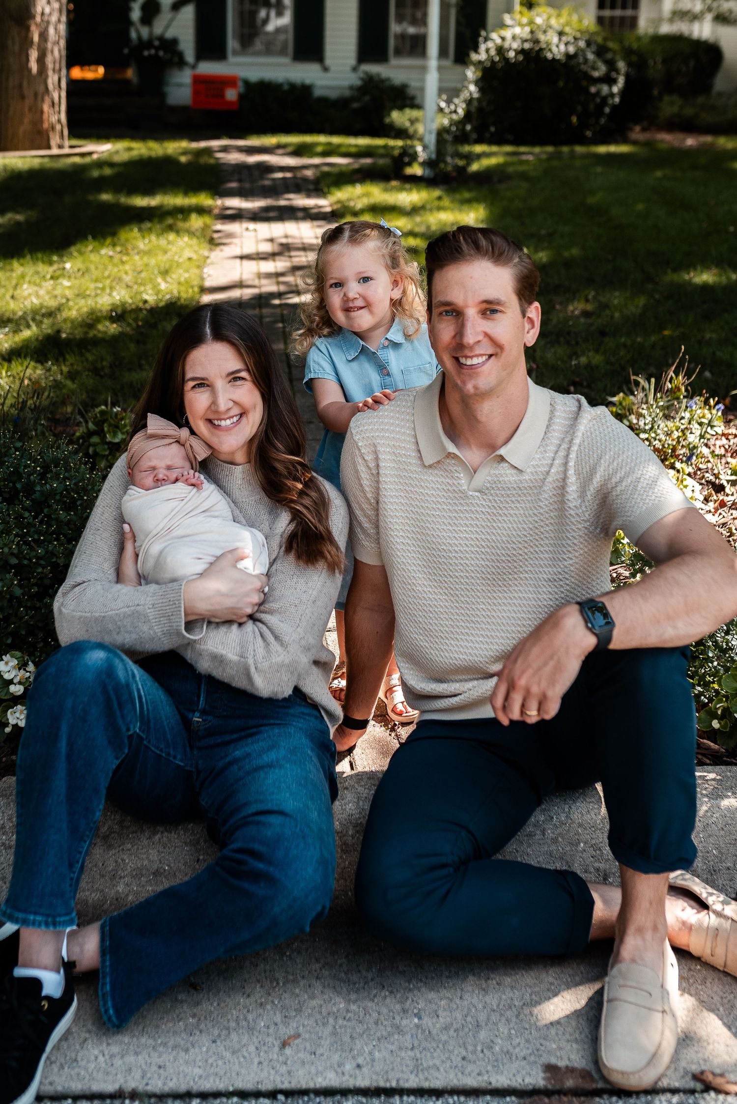 Happy family of four poses outdoors, mom holding newborn, toddler sitting with smiling dad.