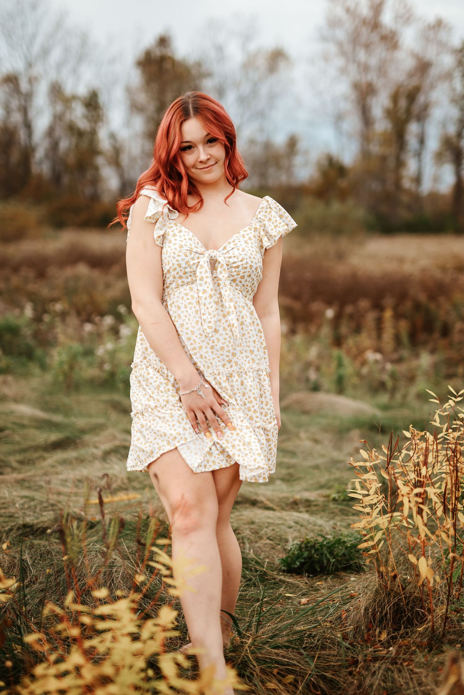 Smiling redhead woman in floral sundress posing in fall meadow with golden plants.
