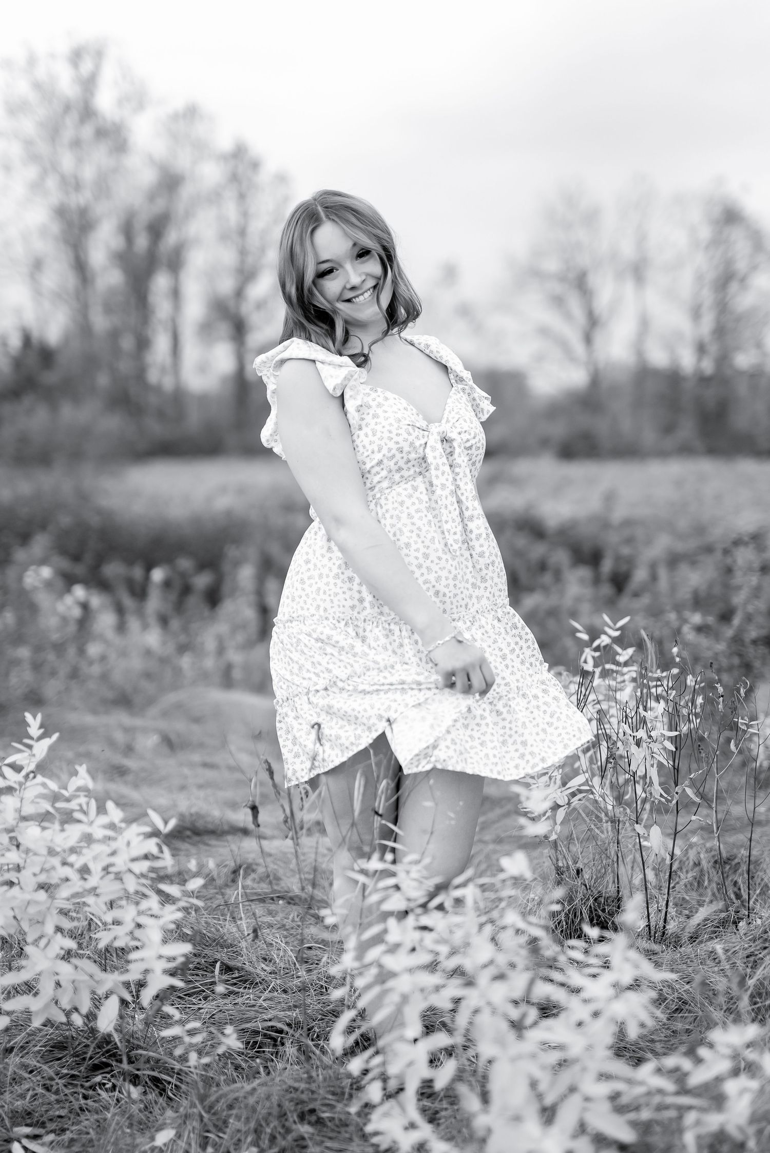 Black and white photo of cheerful young woman in floral dress posing playfully in a meadow.