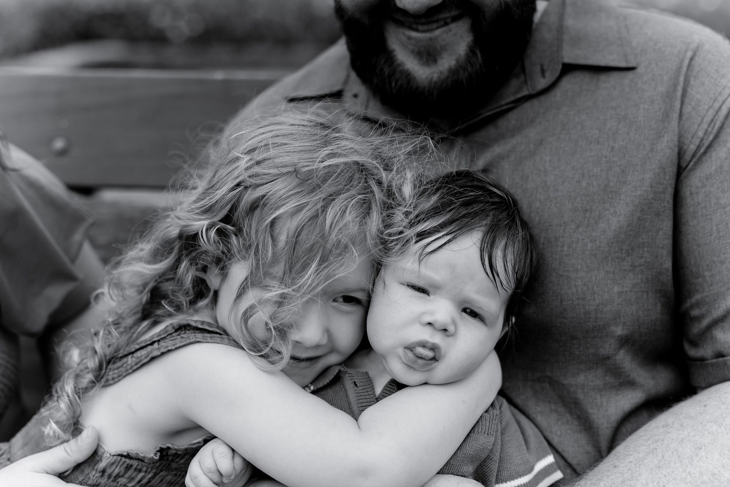 Black and white photo of a bearded father hugging two young children, a toddler and a curly-haired girl, outdoors.