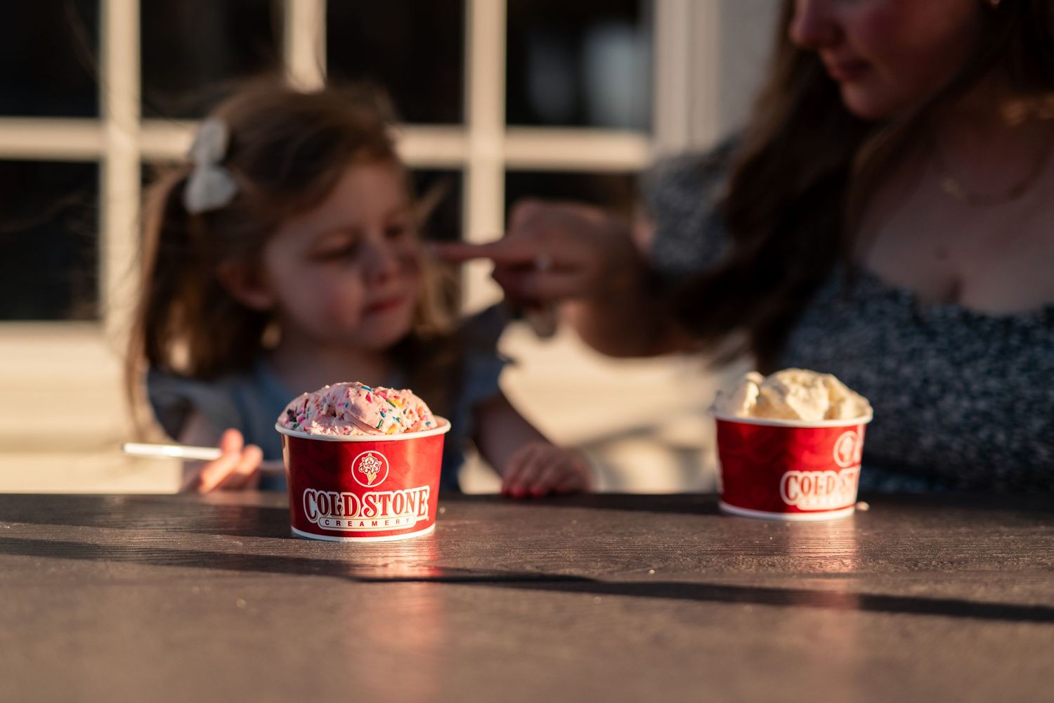 Two Cold Stone Creamery ice cream cups on a table with a woman and young girl enjoying treats outdoors.