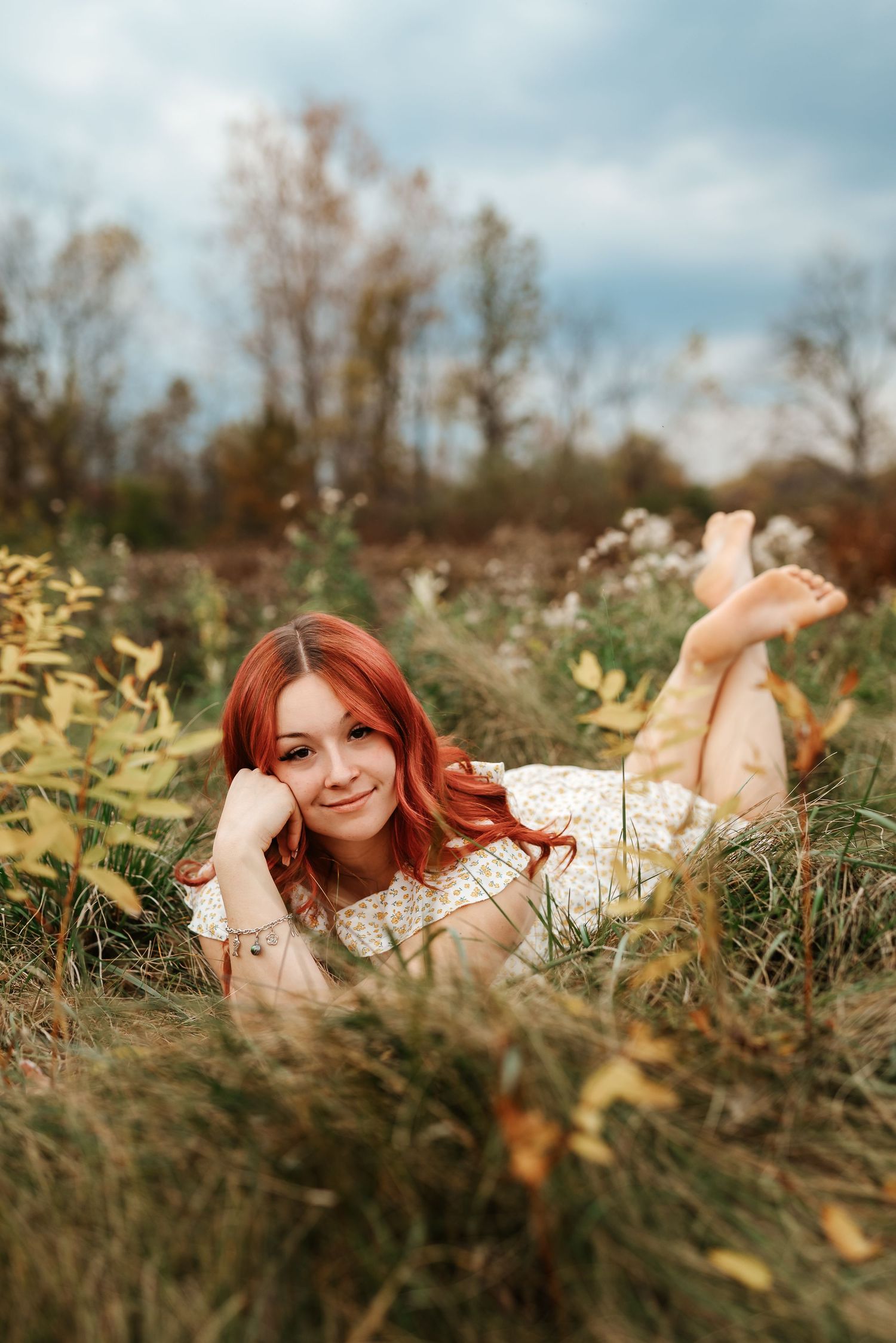 A young redhead woman lying barefoot in a grassy field, smiling at the camera on a cloudy autumn day.