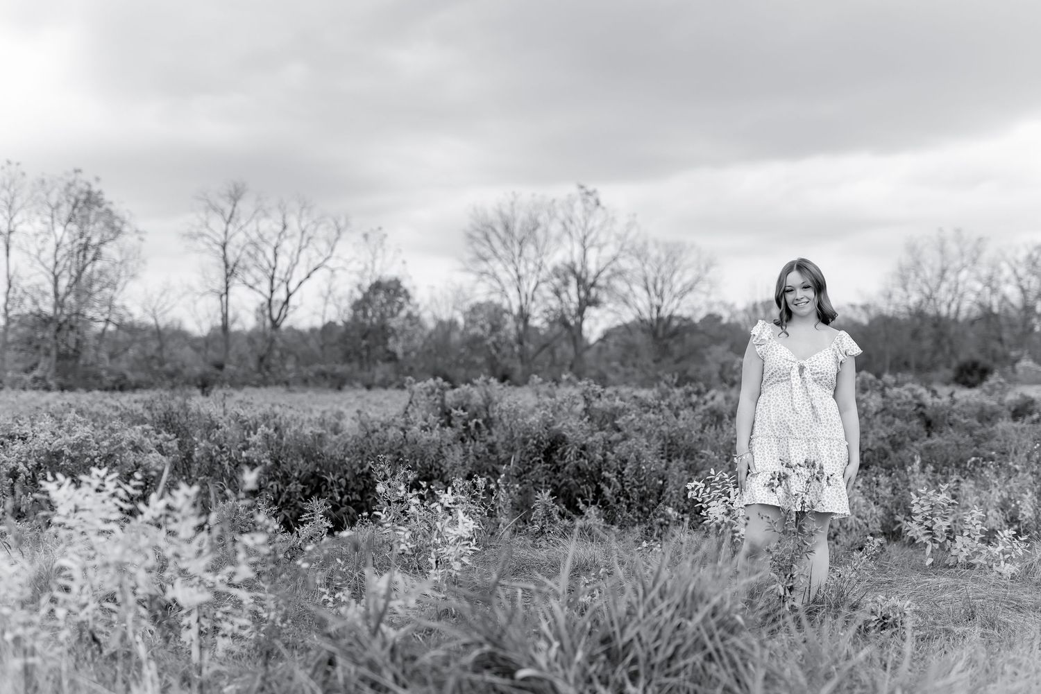 Young woman in floral dress standing in open field on cloudy day, black and white.