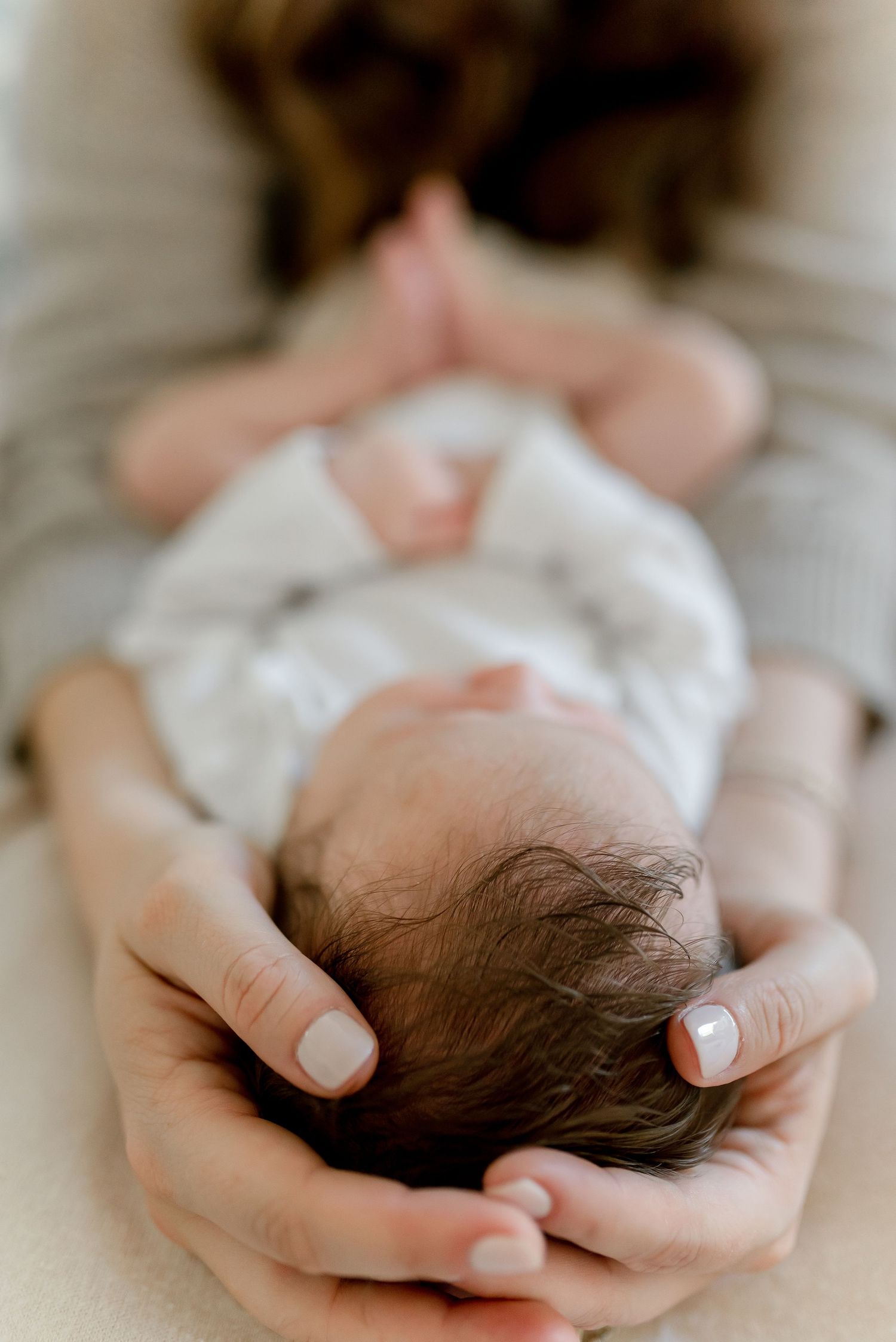 Mother gently cradling newborn baby's head in her hands, close-up of infant with dark hair lying on soft surface.