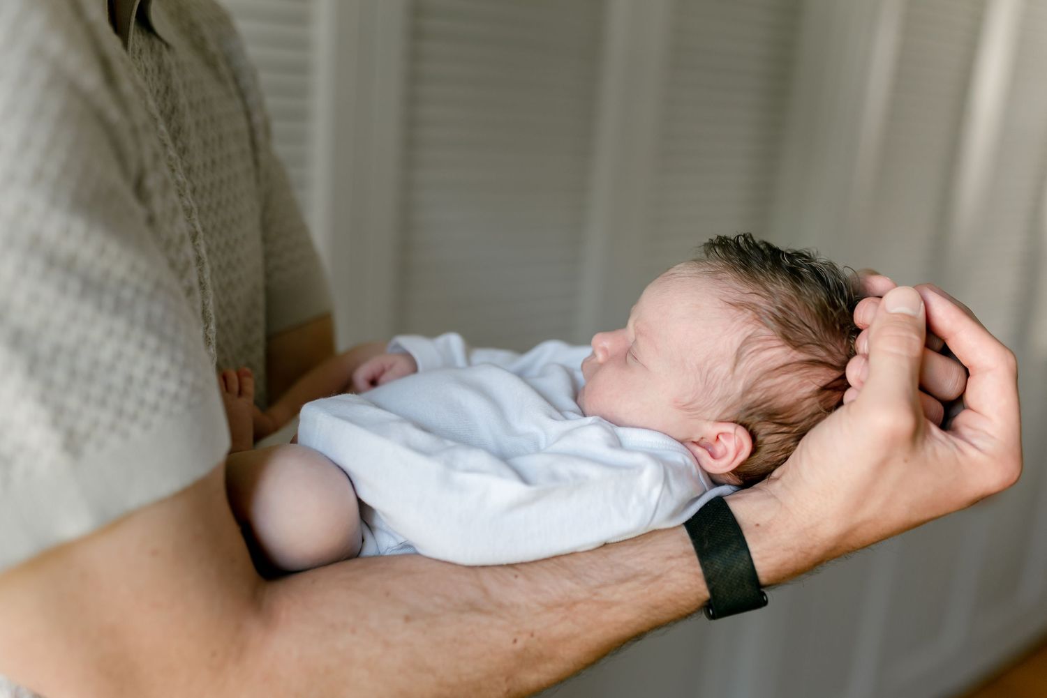 Newborn baby sleeping peacefully in parent's supportive hands, wearing a white onesie.