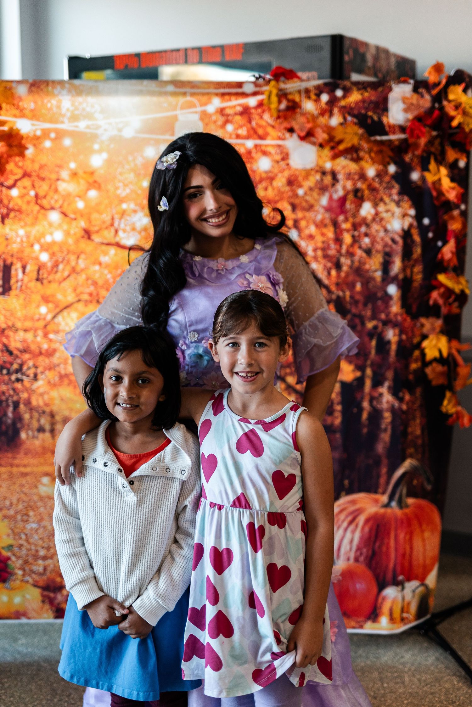 Woman dressed as a princess poses with two young girls in front of an autumn-themed photo backdrop.