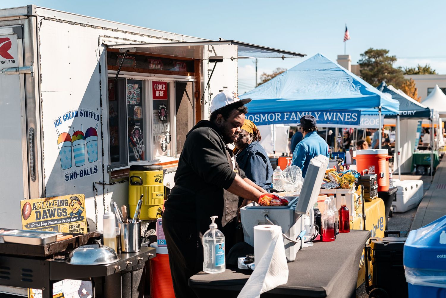 Vendor working at an outdoor food market booth with colorful supplies and a blue tent in the background.