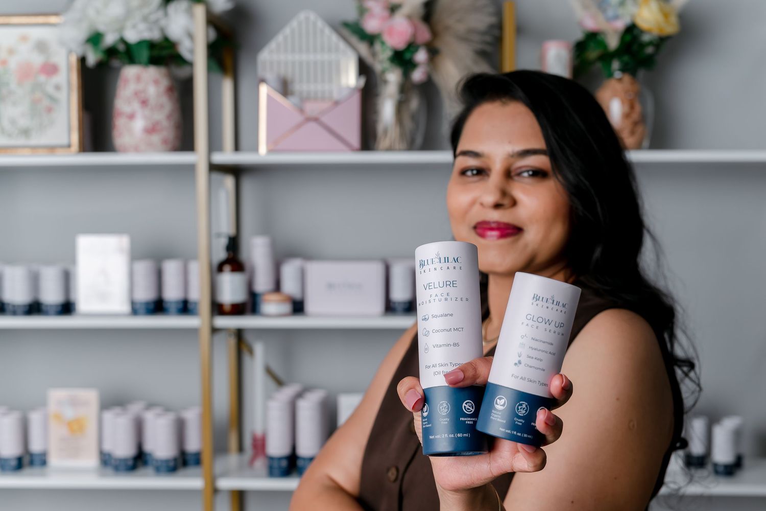 Woman smiling holding two blue skincare product containers in a boutique beauty shop with shelves of products.