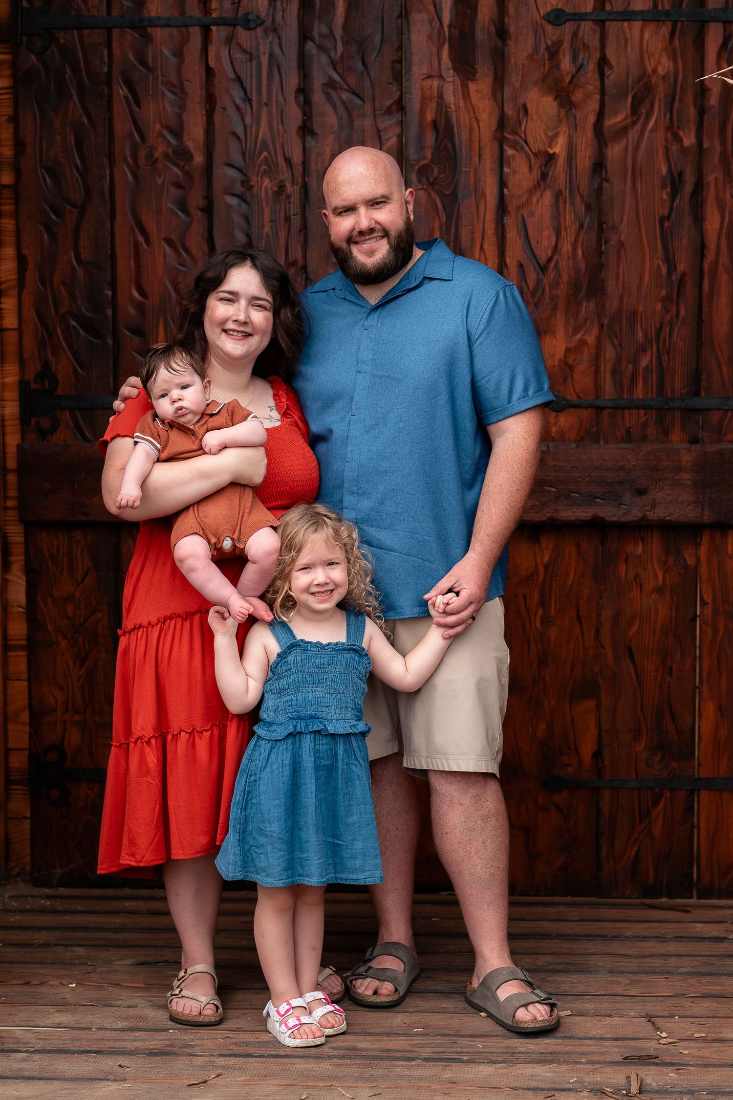 Family of four posing in front of rustic wooden doors, parents with two young children including a baby.