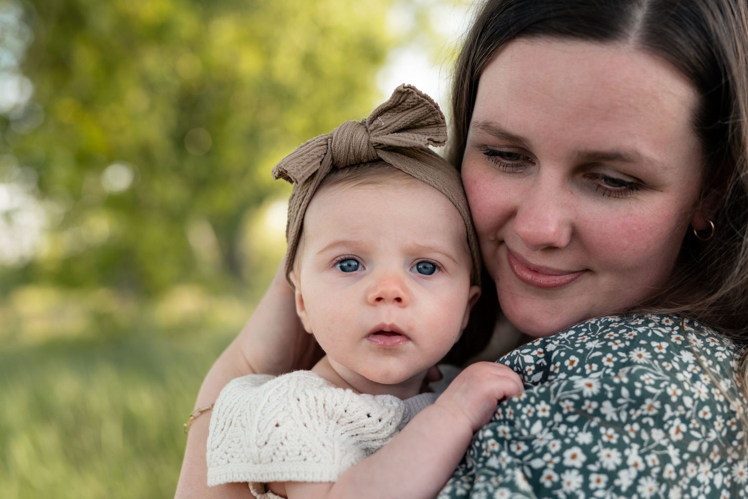 A mother tenderly holds her blue-eyed baby girl wearing a bow headband outdoors in a lush green setting.