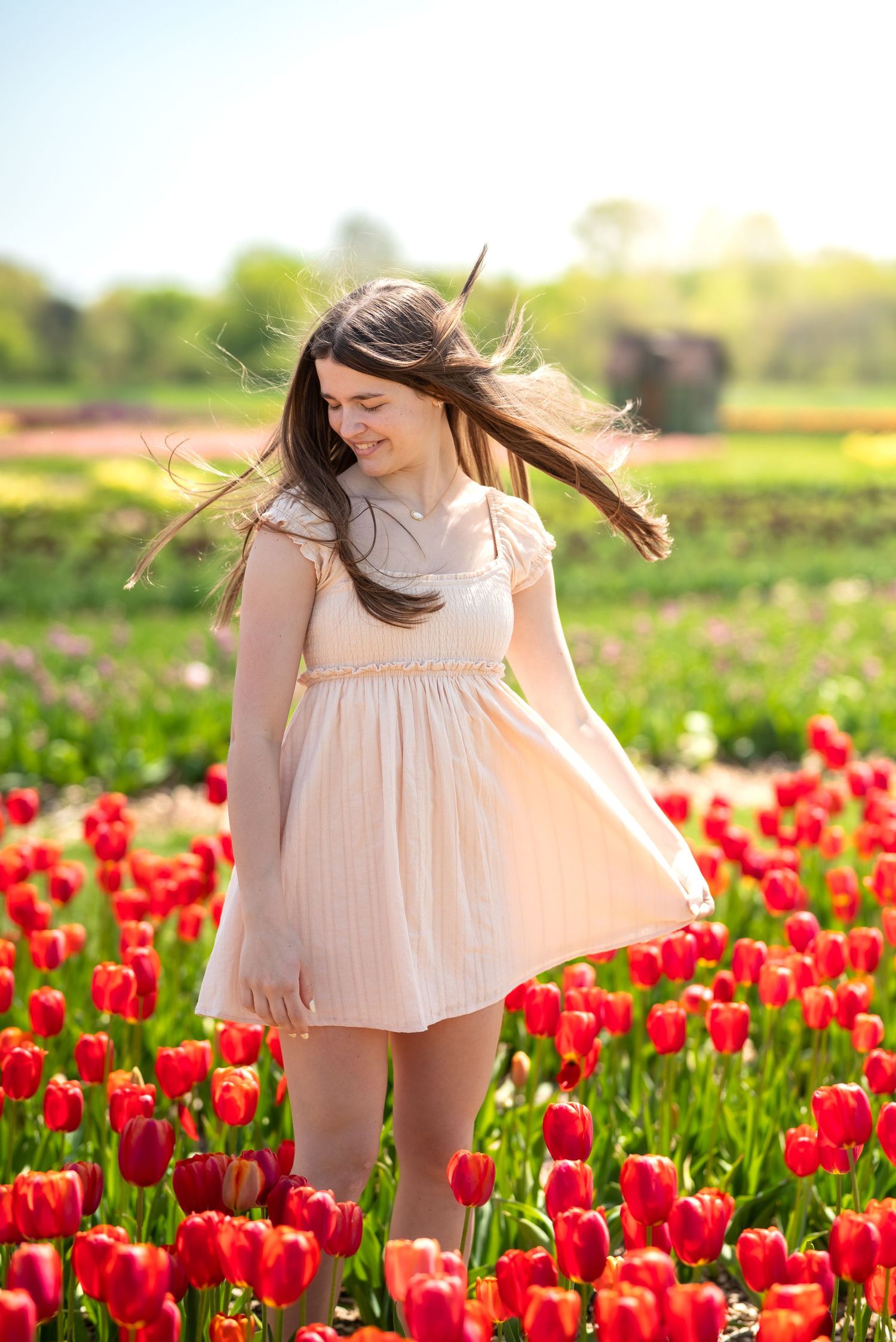 Young woman in white dress standing in vibrant red tulip field, hair blowing in wind on sunny spring day.
