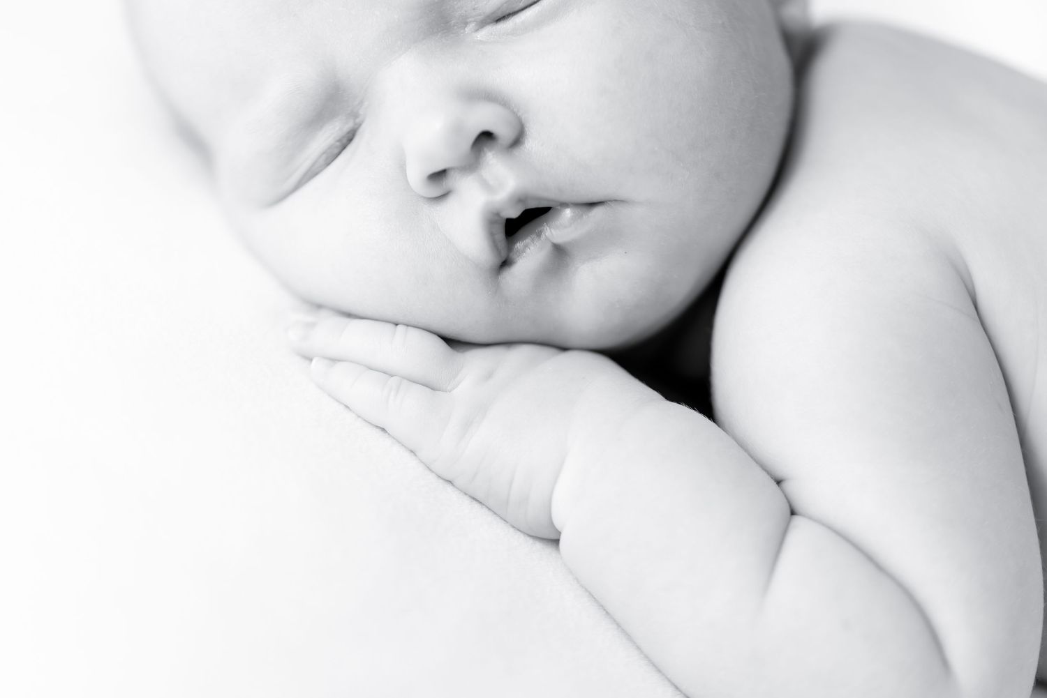 Black and white newborn baby sleeping peacefully with tiny hands tucked under cheek in a serene close-up photo.