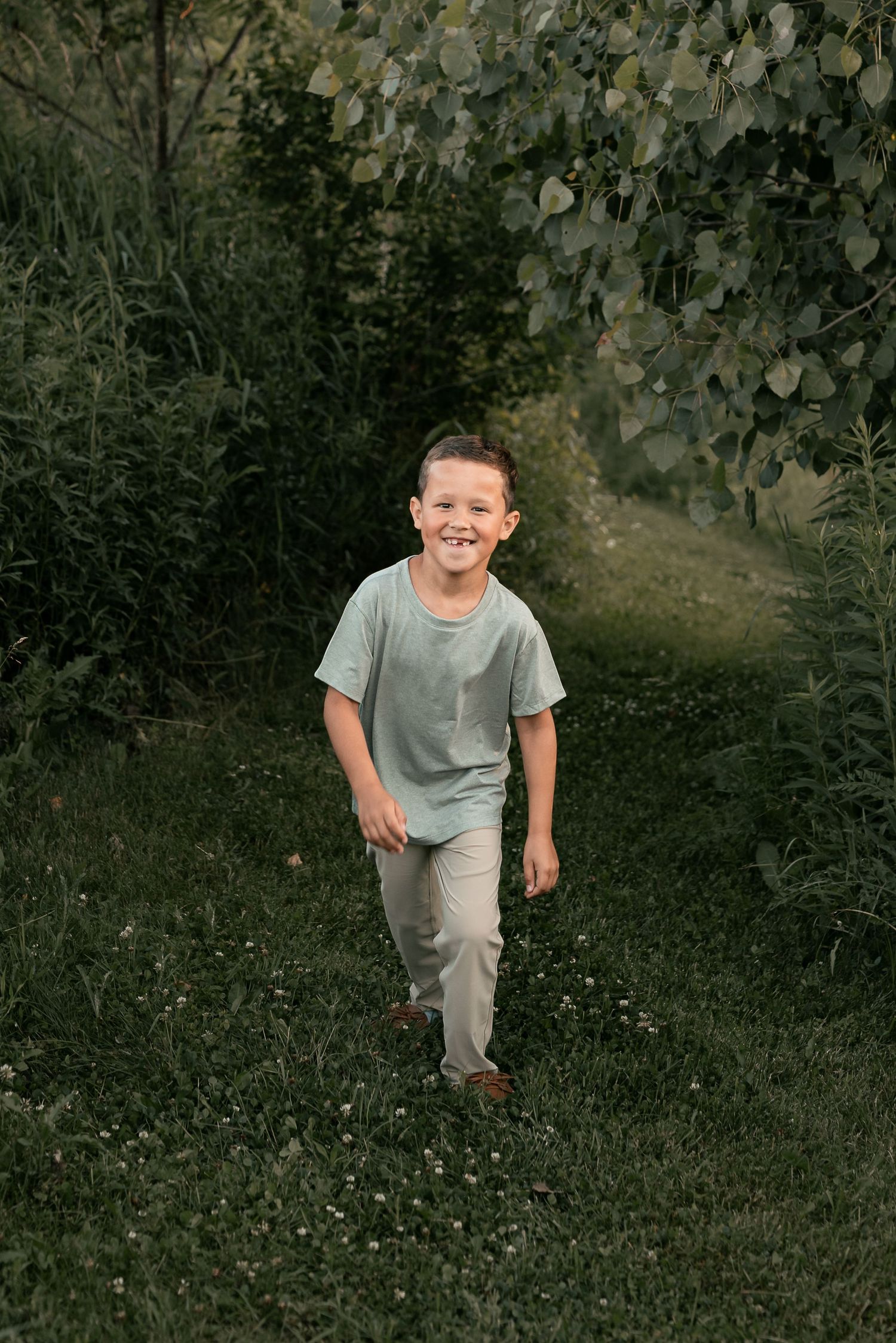 A smiling boy in a mint green t-shirt and khaki pants walks through a lush green garden path.