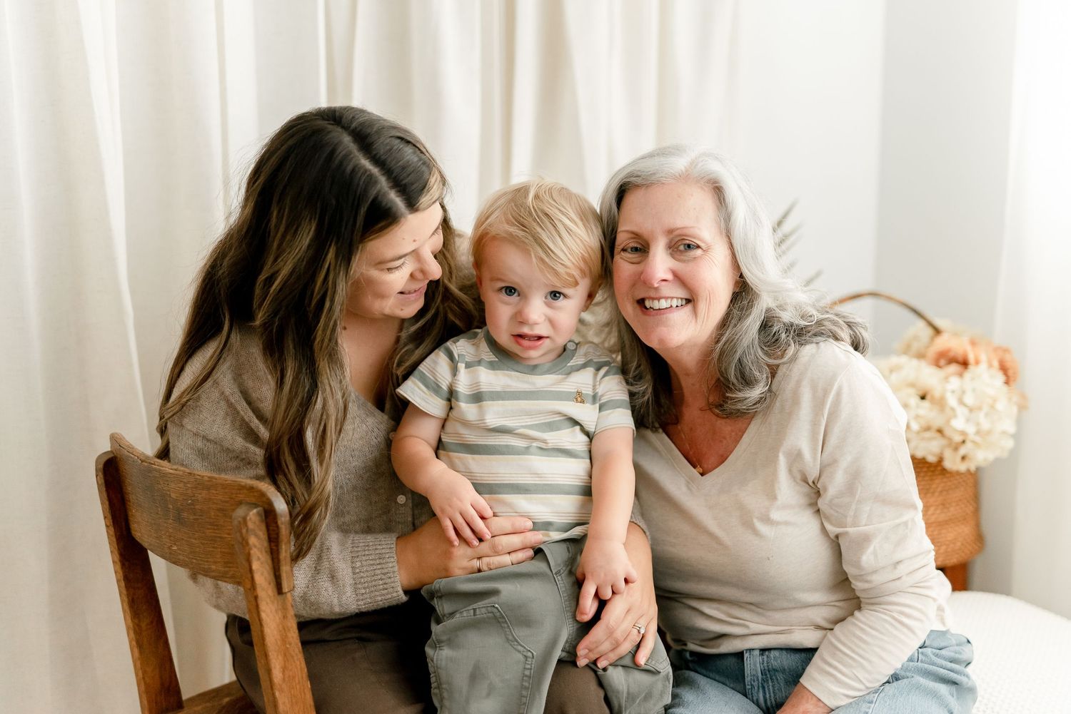 Two women and a toddler boy smile together in a bright studio portrait, capturing a warm three-generation family moment.
