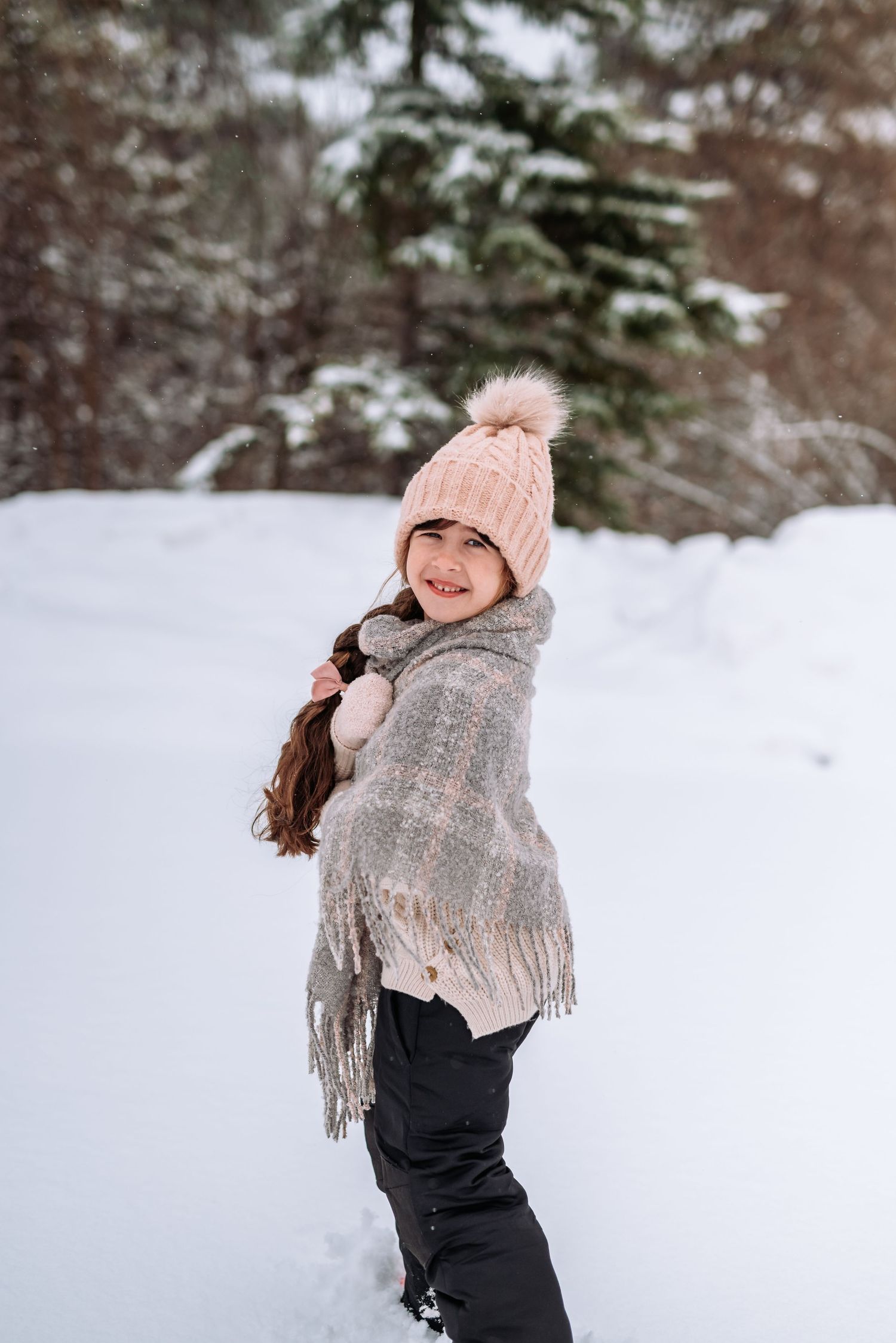 Cozy winter scene with pink beanie, gray scarf, and black outfit against snow-covered trees.
