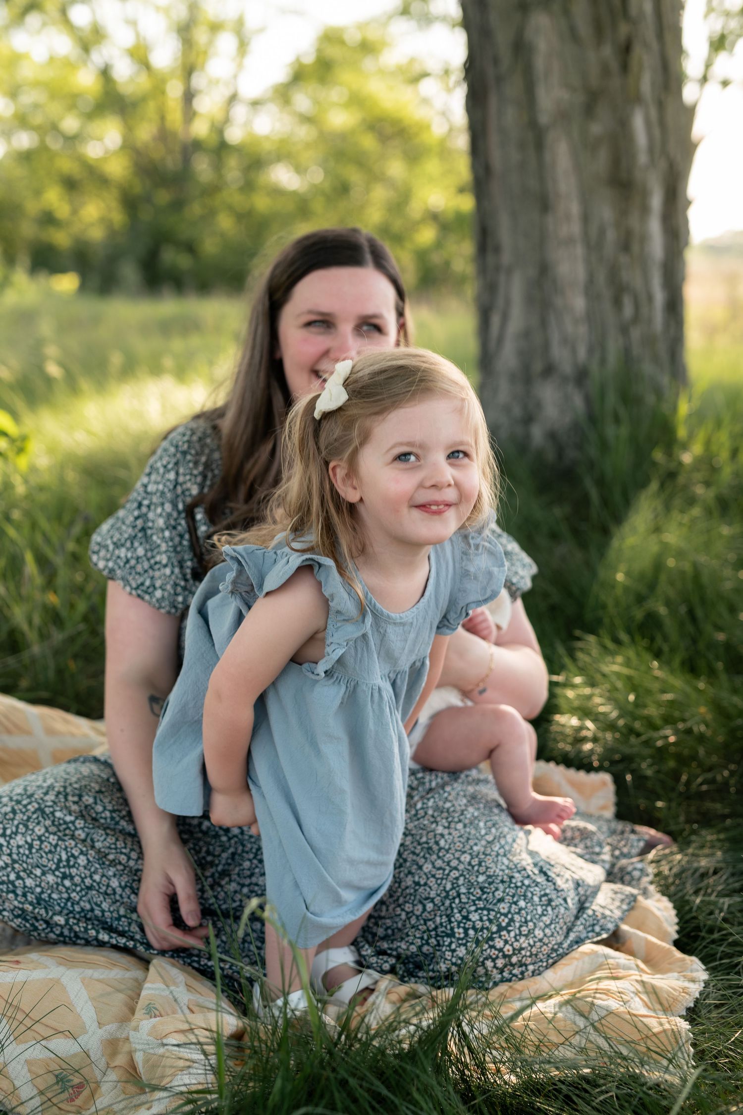 A mother and young daughter in matching blue dresses sit together on a blanket outdoors near a tree in golden sunlight.