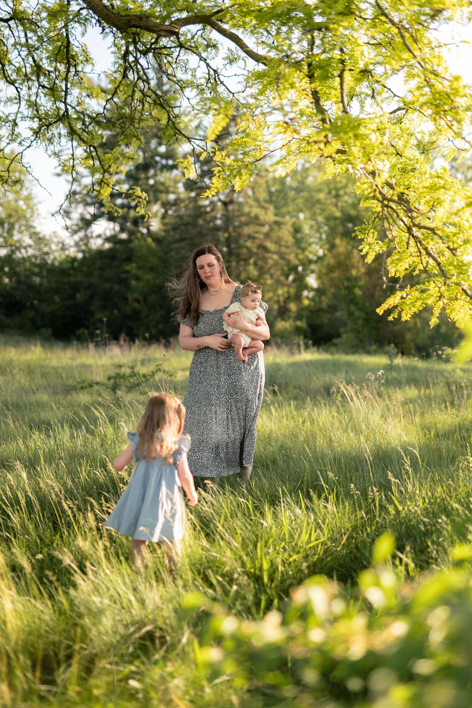 Mother in floral dress holds baby while young girl in blue dress walks through sunlit green meadow.