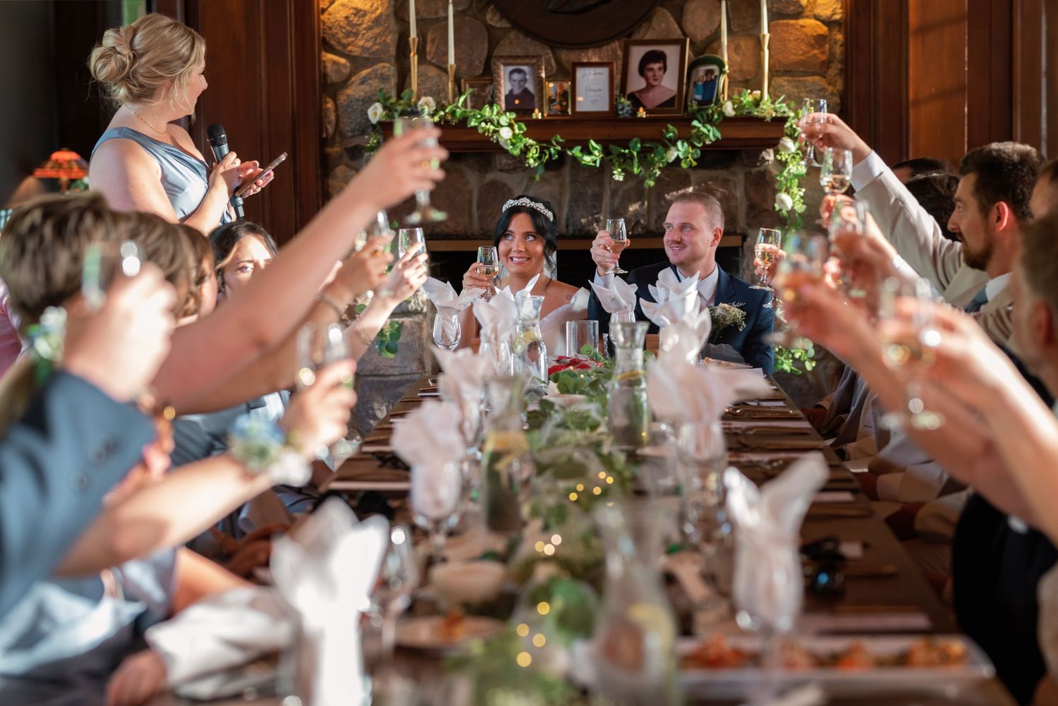 Wedding reception guests raise their glasses in a toast at an elegant wood-paneled dining room decorated with floral garlands.