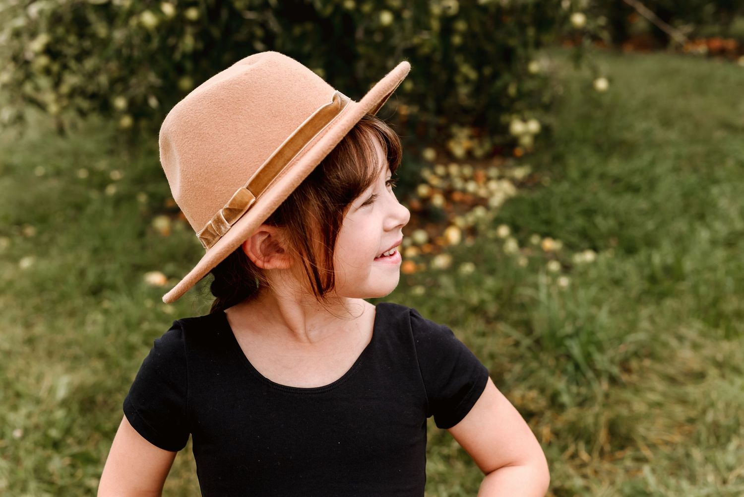 A person wearing a brown hat and black shirt stands in a garden setting with sunlit trees in the background.