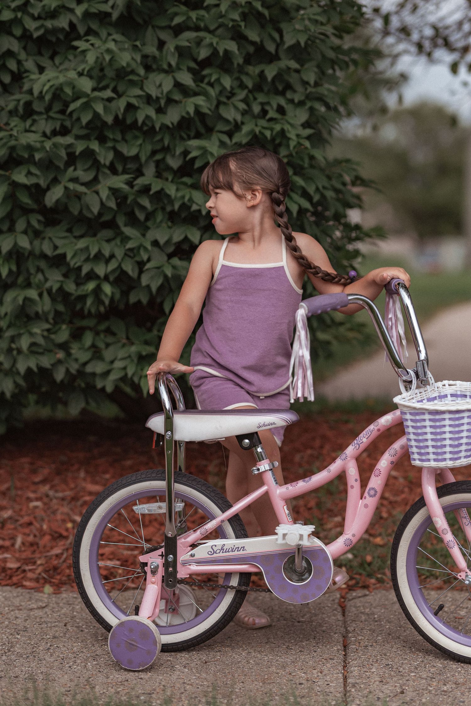 Child seated on pink bicycle with training wheels and decorative basket against green foliage background.