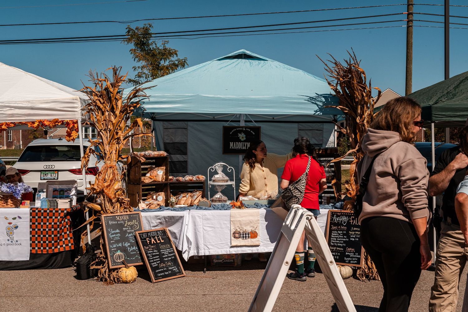 Autumn farmers market with corn stalks, vendor tents, and shoppers browsing baked goods on a sunny fall day.
