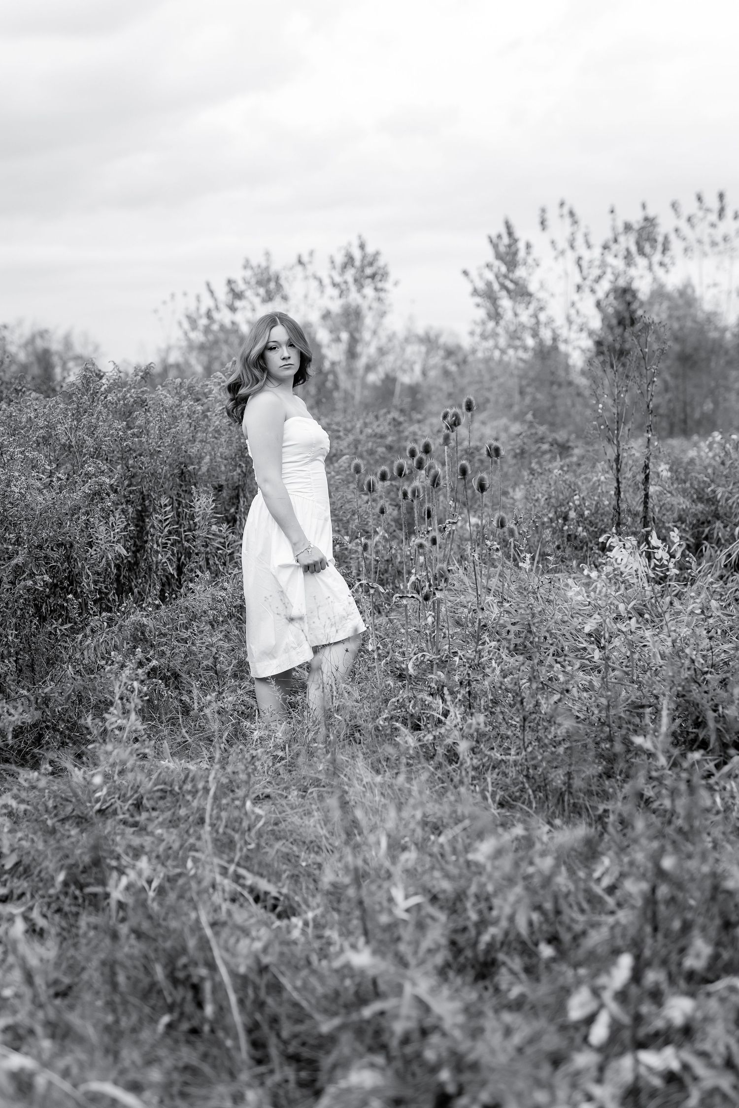 Young woman in white dress standing among wildflowers in a black and white field photo.