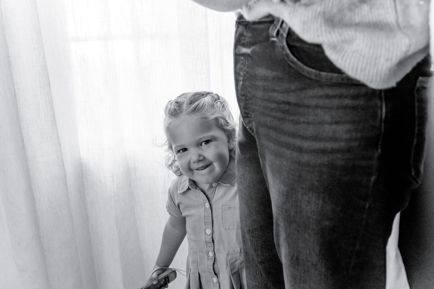 Black and white photo of a smiling toddler girl in a dress standing beside an adult in jeans near a bright window.