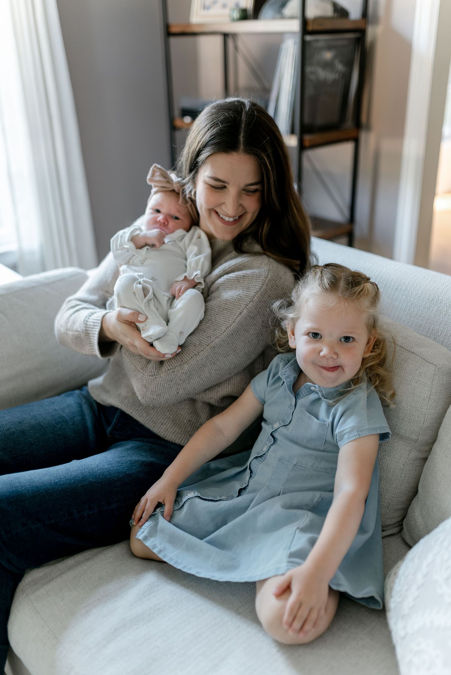 Smiling mother holding newborn baby while toddler girl in blue dress sits beside her on white sofa.