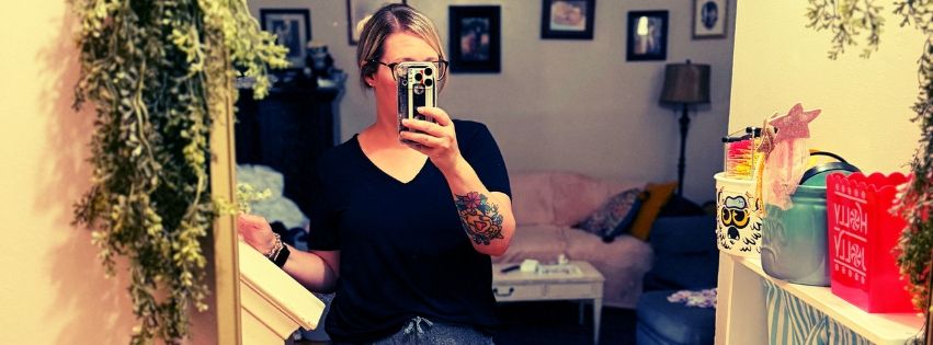 Woman with tattoos taking a mirror selfie in a cozy decorated room with plants and framed pictures.