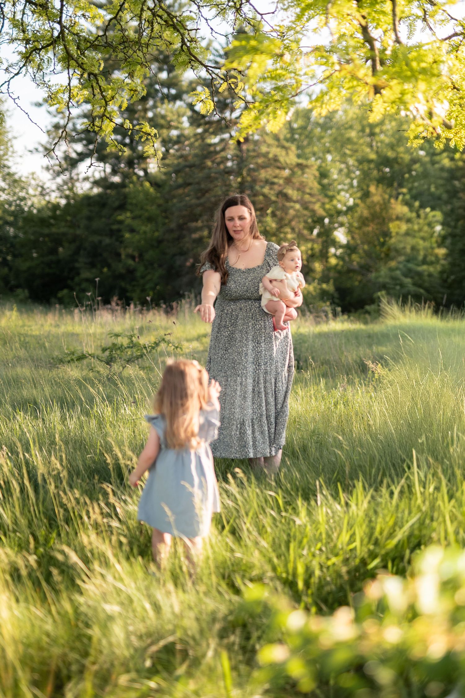 Mother in floral dress holds baby while young girl runs toward her in sunny green meadow.