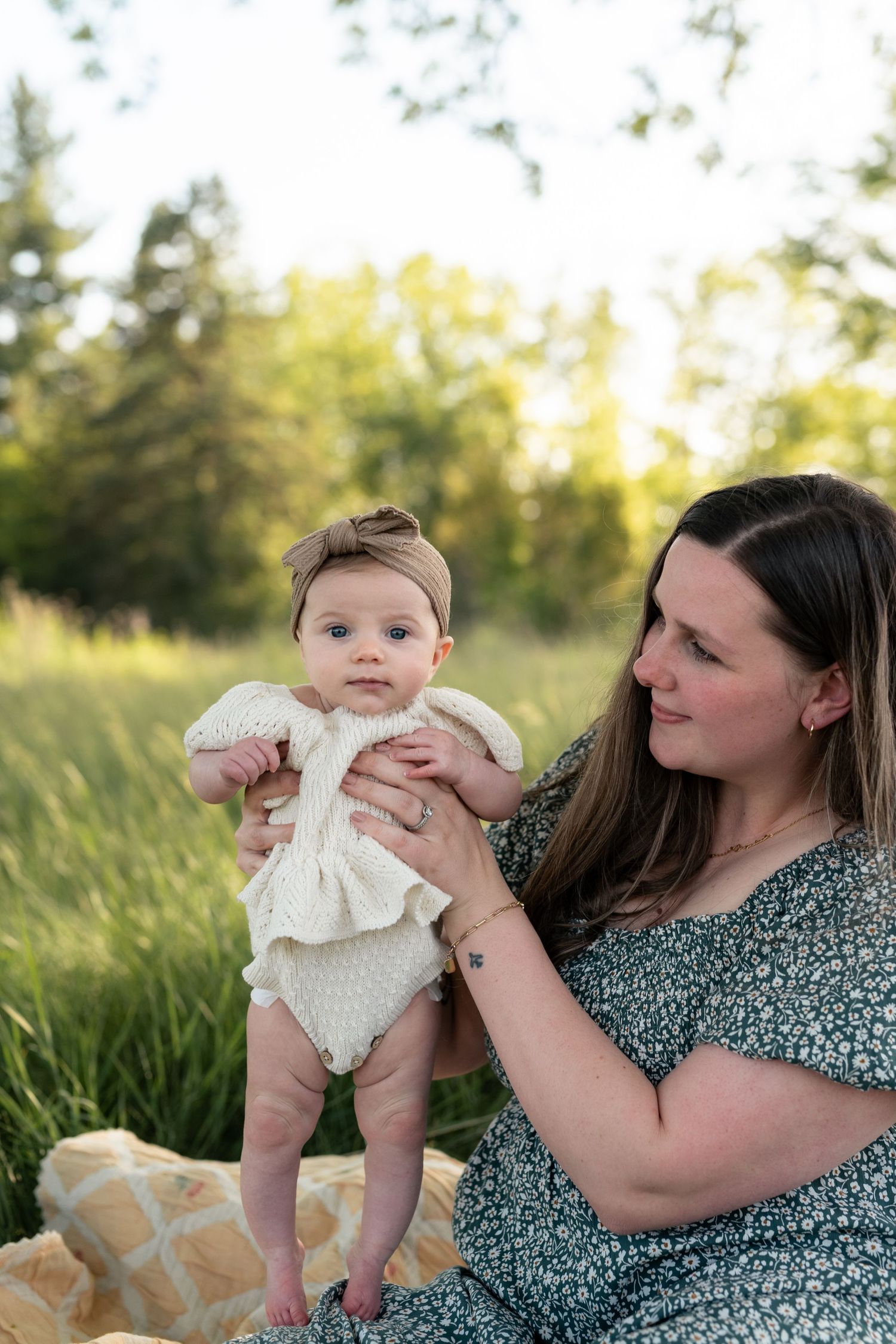 A mother holds her baby girl in a white knit romper on a blanket in a sunny green field.