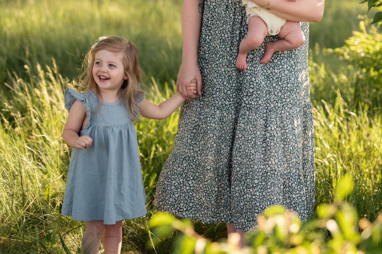 A young girl in a blue dress holds her mother's hand in a sunny green meadow, as mom holds a baby.