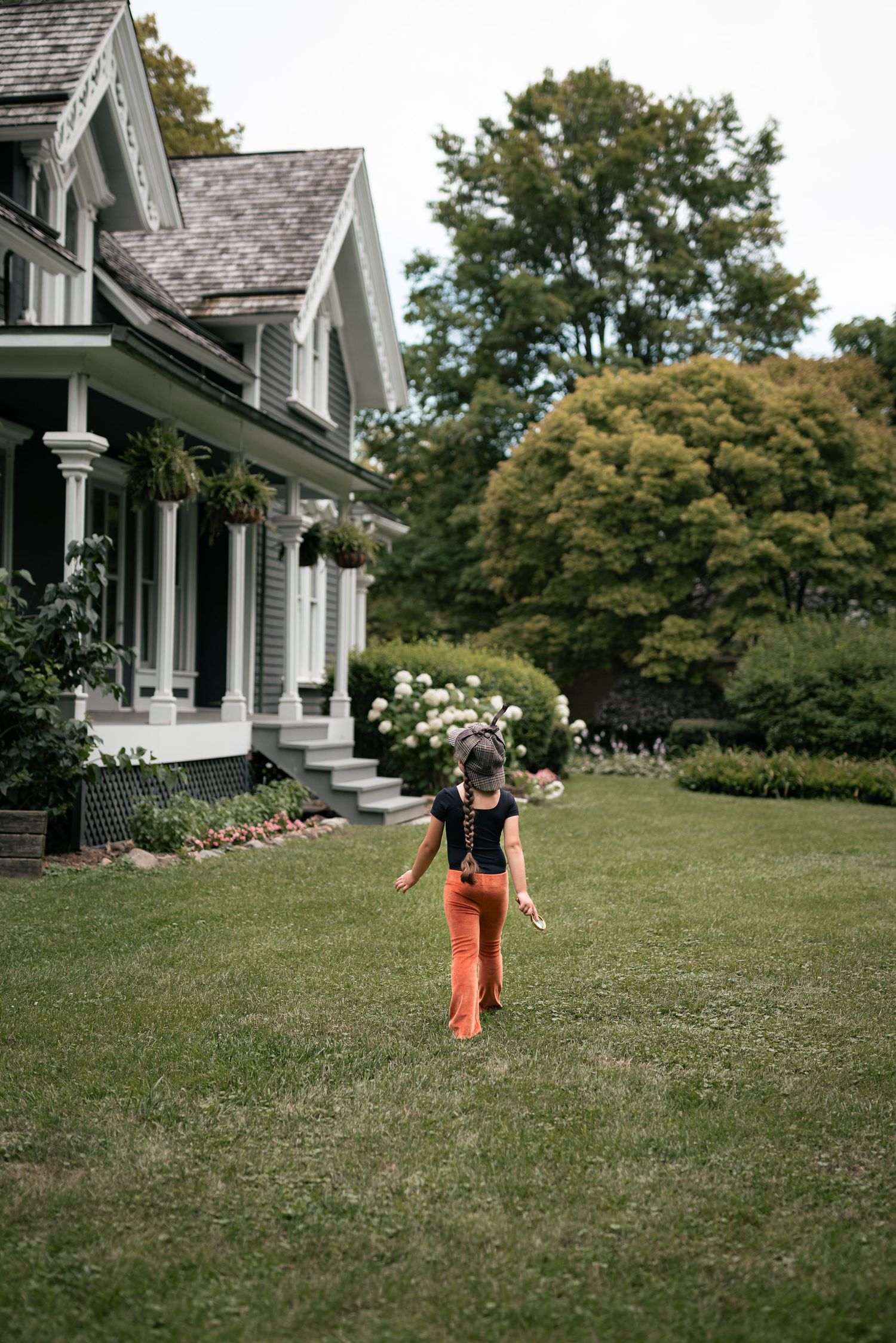 A woman in orange pants and black top walks across a lush green lawn beside a charming gray Victorian-style home.