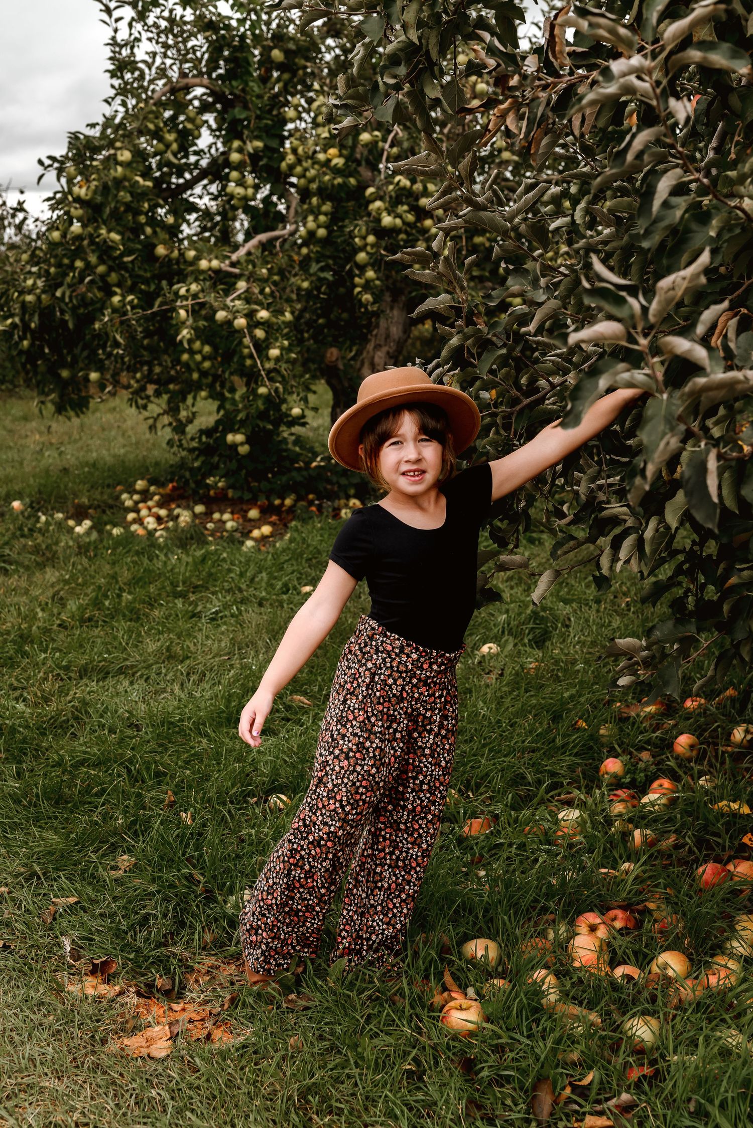 A person in a floral dress dances joyfully in an apple orchard with fallen fruit scattered on the ground.