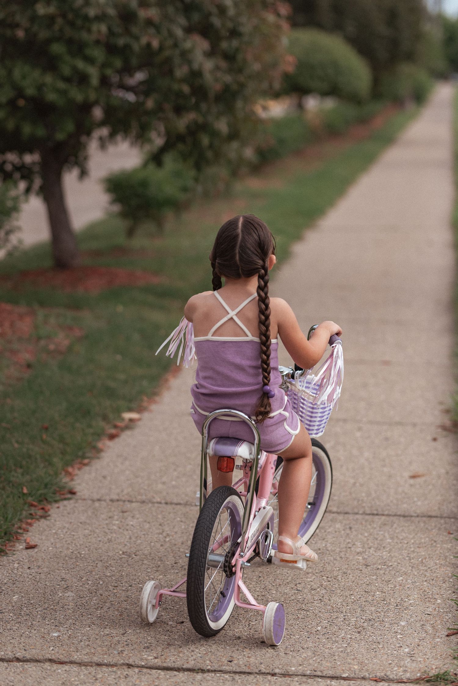 Young cyclist in purple outfit riding training wheels on neighborhood sidewalk lined with trees and grass.