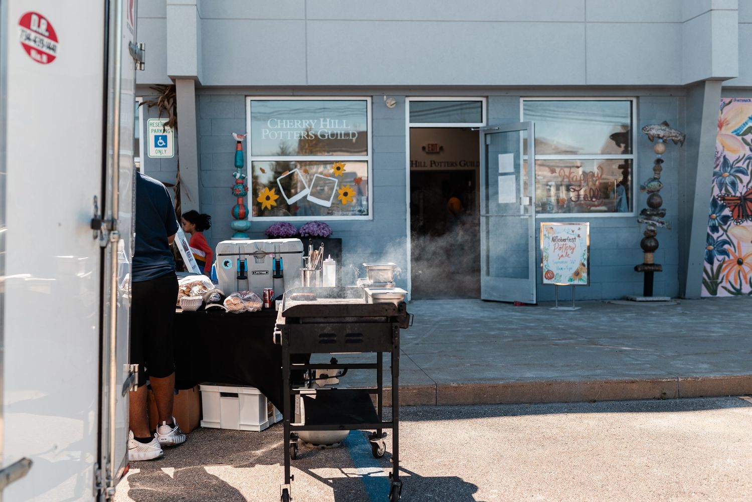 Exterior of a gray storefront with a vendor grilling outside on a sunny day, steam rising from the grill.