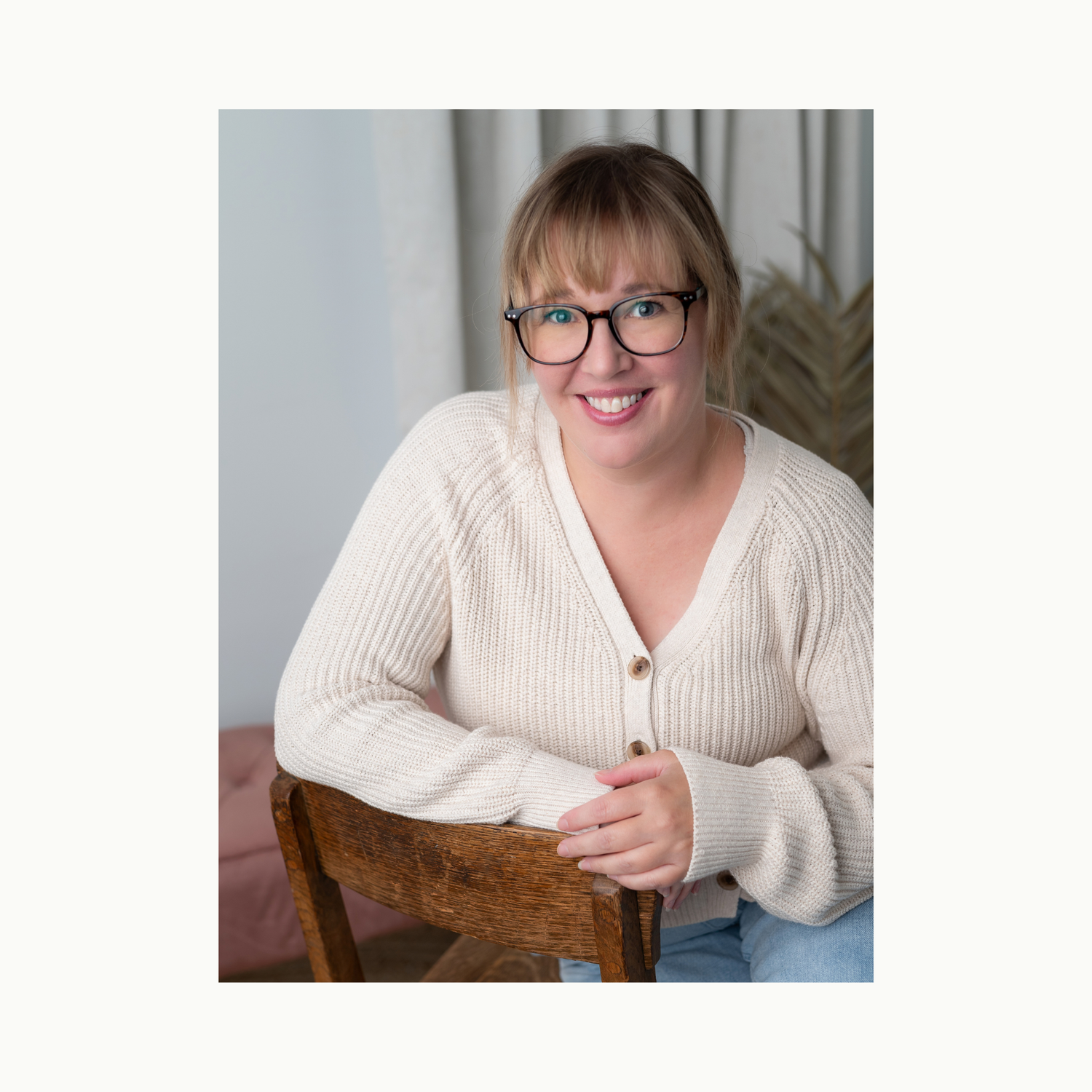 Smiling woman with glasses and bangs wearing a cream cardigan, seated casually on a wooden chair in a bright studio.