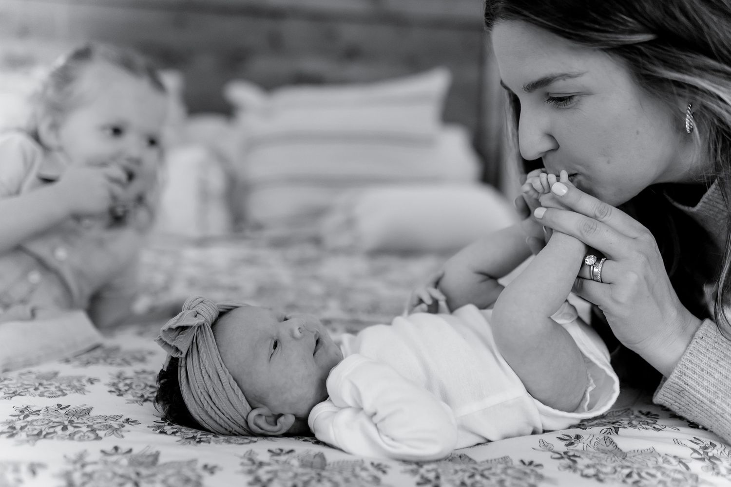 Black and white photo of mother kissing newborn baby's hand while toddler watches on bed.