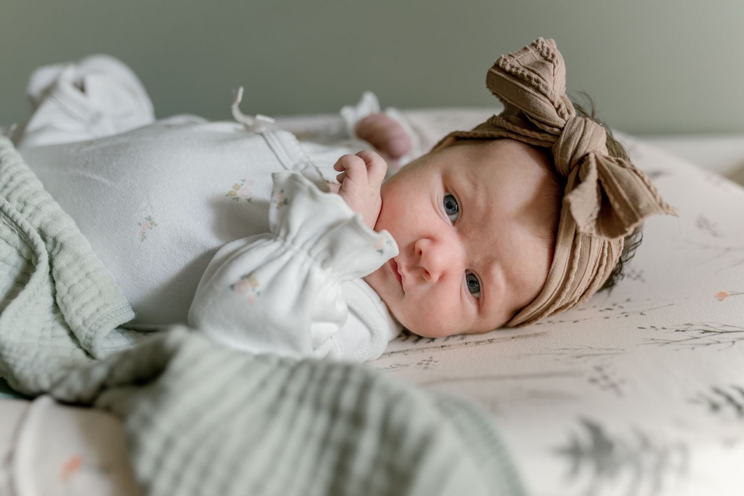 Newborn baby in white outfit and tan bow headband lying on patterned blanket.