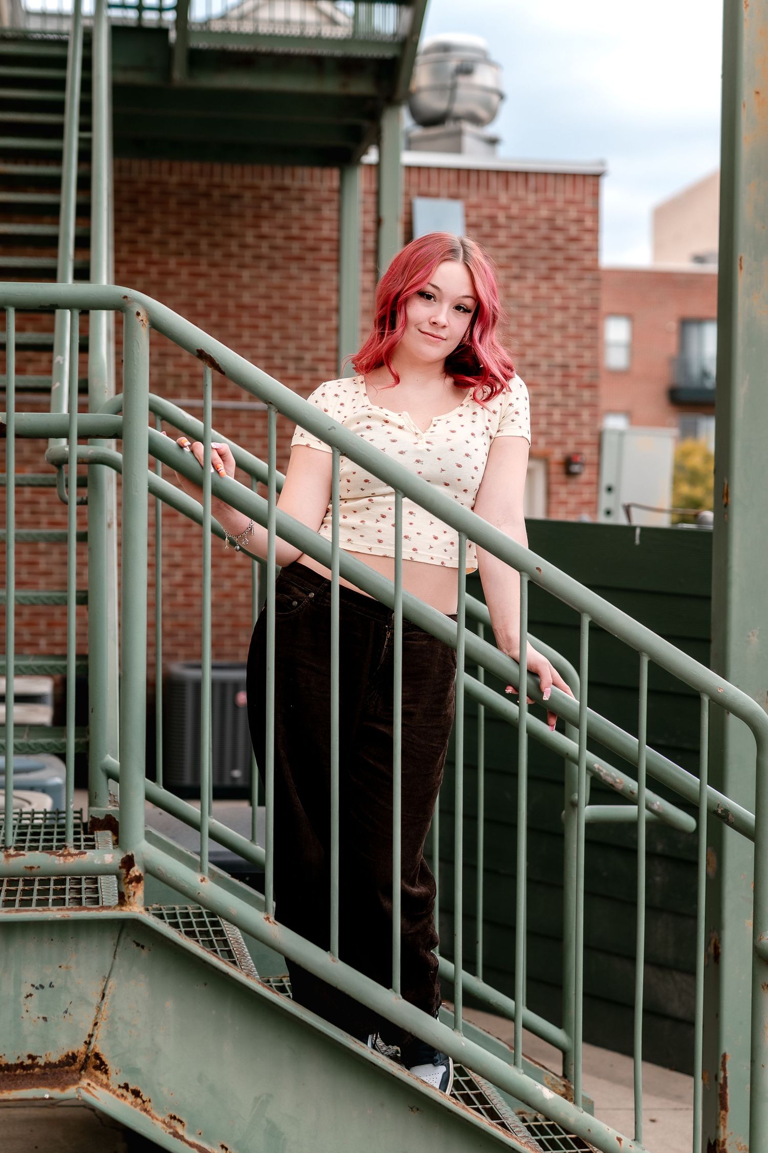 Young woman with red hair poses on green metal fire escape stairs against brick building backdrop.